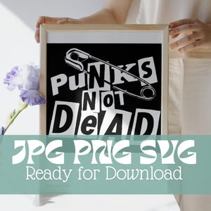 May include: Black and white framed art print with the text "Punks Not Dead" and a safety pin graphic. The image also includes the text "JPG PNG SVG Ready for Download". The artwork is held by a person.
