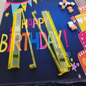 Puede incluir: Soportes de fichas de mahjong de pl&aacute;stico amarillo y fichas sobre una superficie azul oscuro con las palabras "HAPPY BIRTHDAY!" en letras coloridas. El fondo incluye ilustraciones de pasteles de cumplea&ntilde;os y elementos decorativos.