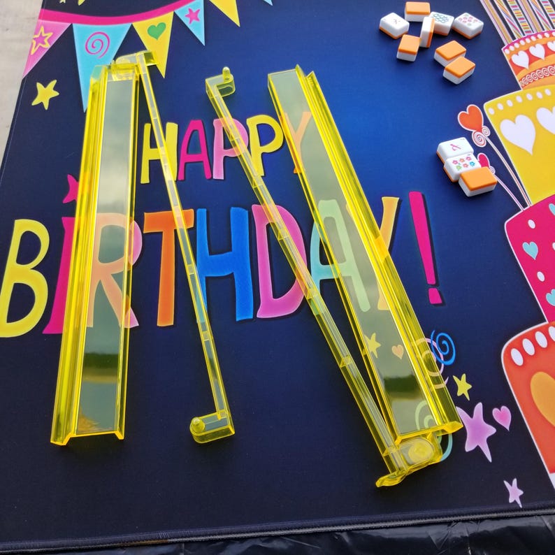 Puede incluir: Soportes de fichas de mahjong de pl&aacute;stico amarillo y fichas sobre una superficie azul oscuro con las palabras "HAPPY BIRTHDAY!" en letras coloridas. El fondo incluye ilustraciones de pasteles de cumplea&ntilde;os y elementos decorativos.