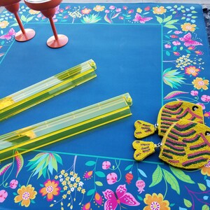Puede incluir: Un mantel azul con un borde floral con mariposas y flores. Dos bandejas de pl&aacute;stico amarillas y dos monederos amarillos con forma de pez con cuentas est&aacute;n sobre el mantel. Dos copas de vino rojas est&aacute;n en la esquina superior izquierda.