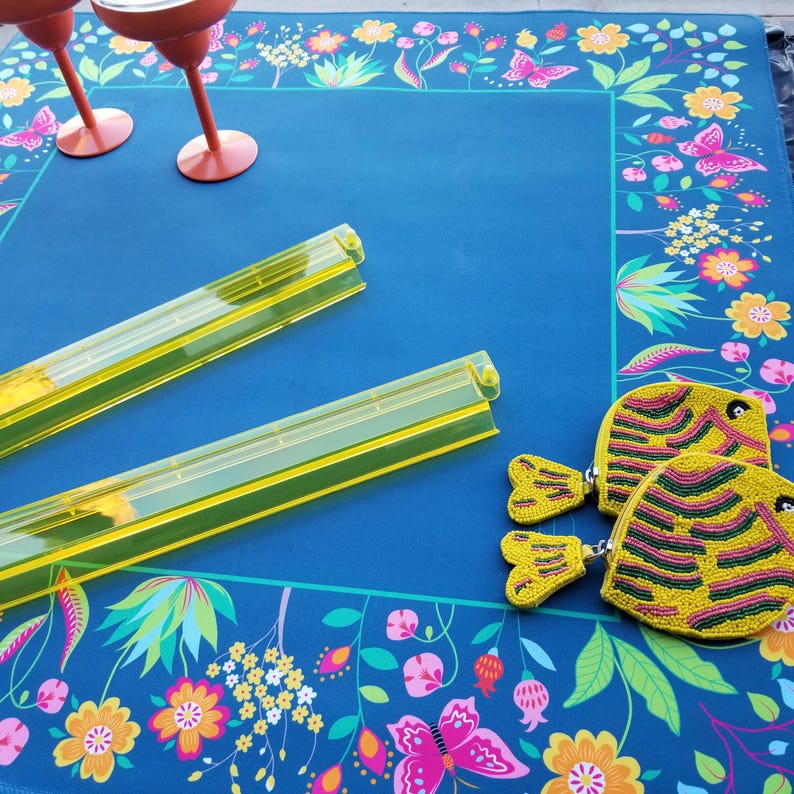 Puede incluir: Un mantel azul con un borde floral con mariposas y flores. Dos bandejas de pl&aacute;stico amarillas y dos monederos amarillos con forma de pez con cuentas est&aacute;n sobre el mantel. Dos copas de vino rojas est&aacute;n en la esquina superior izquierda.