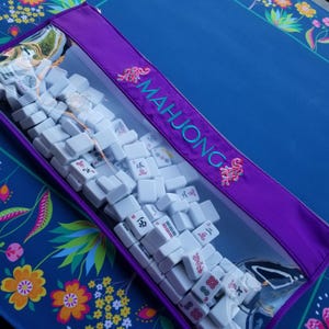 May include: A clear zippered case filled with white Mahjong tiles. The purple case has the word "MAHJONG" embroidered in teal, with pink floral accents. The case is on a blue surface with floral patterns.
