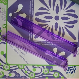 May include: Two transparent purple plastic guides are positioned diagonally across a patterned surface. The surface features a floral design in white and purple, with text that reads "Charleston" and "Passing Options: 1-3: Ceramic Tiles."