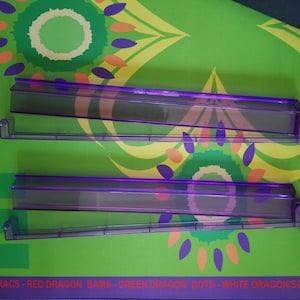 May include: Two transparent purple plastic shelves with a rectangular shape are displayed against a green background with a floral pattern in yellow, orange, and purple. The shelves have a lip on one side and are designed to hold items.