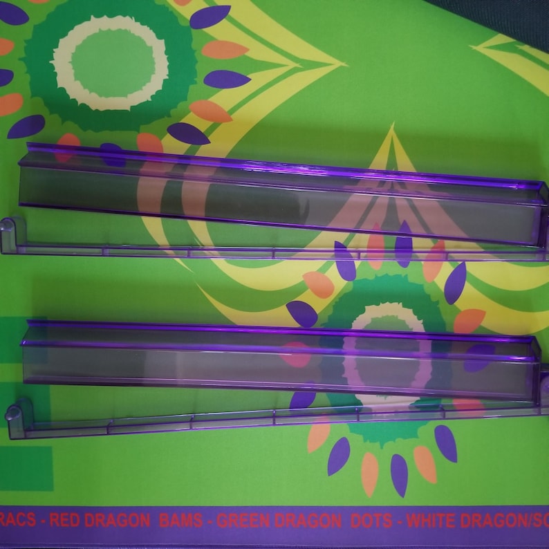 May include: Two transparent purple plastic shelves with a rectangular shape are displayed against a green background with a floral pattern in yellow, orange, and purple. The shelves have a lip on one side and are designed to hold items.