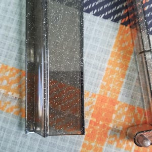 May include: A clear, rectangular object with a glittery, speckled design. The object is a dark gray color and has a reflective surface. The image shows a close-up of the object, highlighting its texture and design. The object is likely a decorative item or part of a larger product.