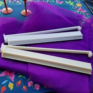 May include: Two white and off-white plastic bag sealers are displayed on a purple cloth. The sealers are rectangular with a long, thin arm extending from one end. The background includes a floral patterned surface and two pink stemmed glasses.