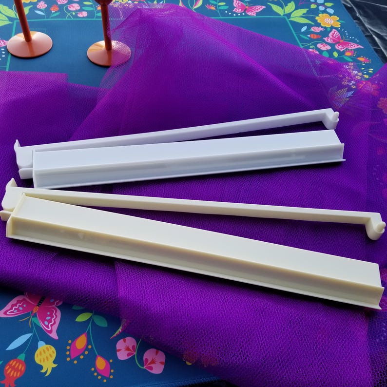 K&ouml;nnte beinhalten: Zwei wei&szlig;e und cremefarbene Plastikbeutelverschl&uuml;sse werden auf einem lila Stoff gezeigt. Die Verschl&uuml;sse sind rechteckig mit einem langen, d&uuml;nnen Arm, der sich von einem Ende erstreckt. Der Hintergrund umfasst eine Oberfl&auml;che mit Blumenmuster und zwei rosa Weingl&auml;ser.