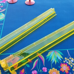Puede incluir: Dos reglas de pl&aacute;stico amarillo transparente con interior verde est&aacute;n colocadas en diagonal sobre una superficie azul. La superficie tiene un estampado floral en la esquina. Dos copas de vino rosas est&aacute;n en el fondo.