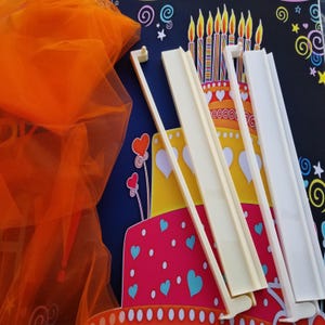 K&ouml;nnte beinhalten: Auf der linken Seite des Bildes ist ein orangefarbener T&uuml;llstoff drapiert. Mehrere cremefarbene Kuchenst&uuml;tzen aus Kunststoff sind vor einer bunten Geburtstagskuchen-Illustration mit Herzen und Kerzen angeordnet.