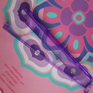 May include: Two transparent purple plastic rulers are positioned diagonally across a pink background with a floral pattern in shades of purple, blue, and teal. The text "CHARLESTON" and other instructions are visible on the lower left.