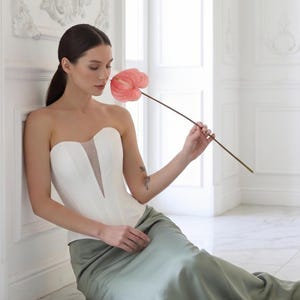 May include: A white strapless corset with a sheer panel and a sage green satin skirt. The model is holding a pink anthurium flower. The setting is a bright room with white walls and marble flooring.