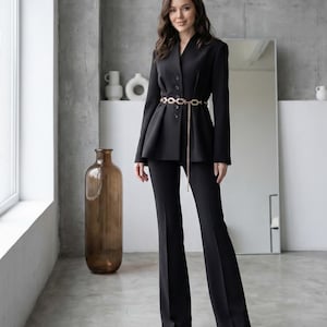 May include: A black suit with a tailored blazer and flared pants. The blazer has a V-neck and is cinched at the waist with a gold chain belt. The pants are long and flowing. The outfit is worn in a modern, minimalist setting.