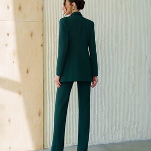 May include: A dark green pantsuit featuring a blazer and matching trousers. The blazer has a classic lapel and a tailored fit. The trousers are straight-legged. The outfit is worn by a person with their back to the camera.