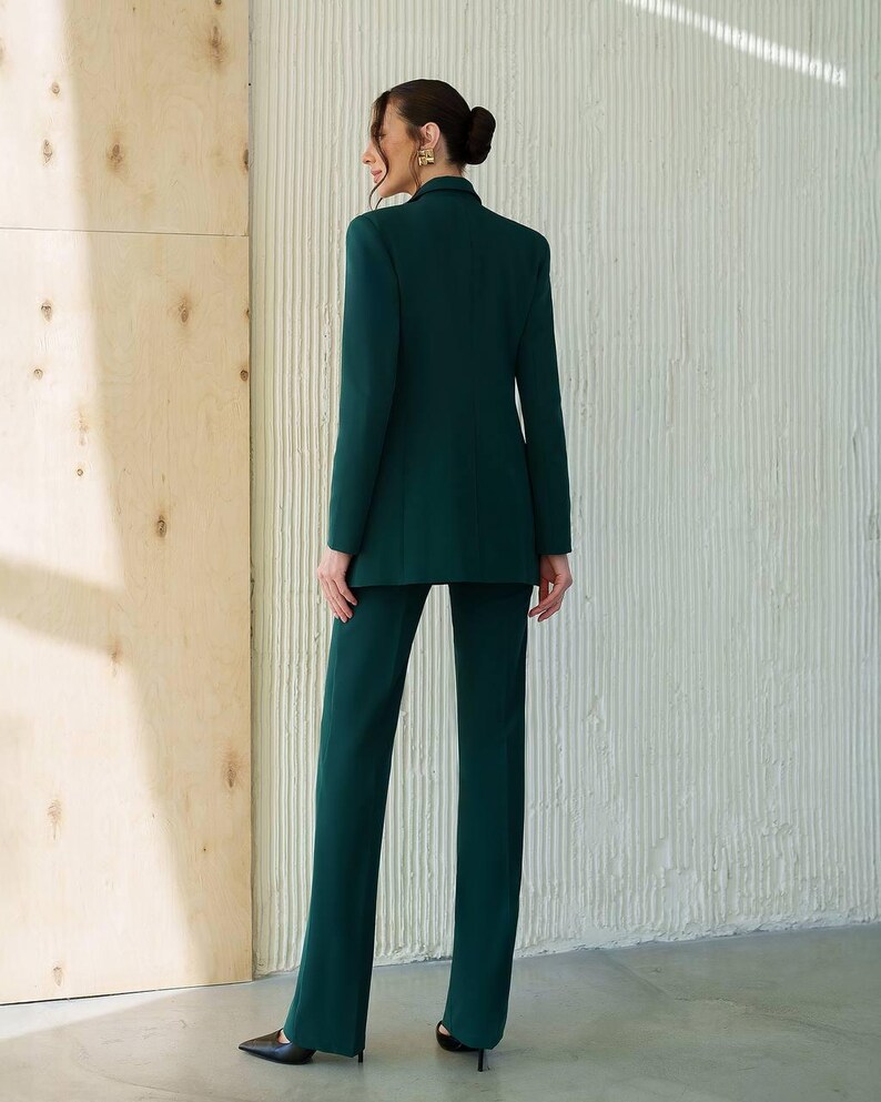 May include: A dark green pantsuit featuring a blazer and matching trousers. The blazer has a classic lapel and a tailored fit. The trousers are straight-legged. The outfit is worn by a person with their back to the camera.