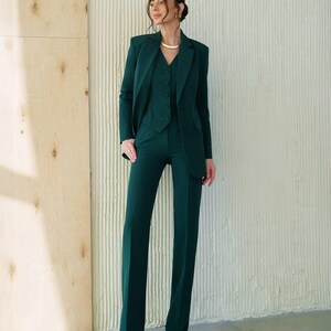 May include: A three-piece emerald green suit. The suit includes a blazer, vest, and wide-leg trousers. The blazer has a notched lapel and the vest is buttoned. The trousers are long and flared. The suit is made of a smooth fabric.