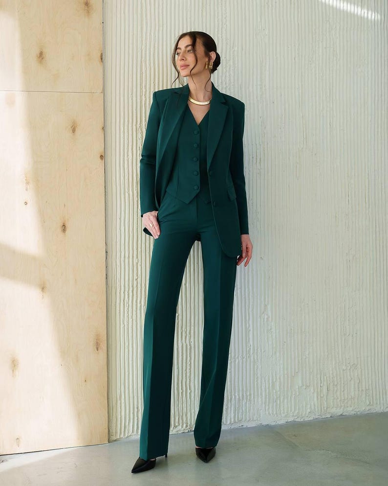 May include: A three-piece emerald green suit. The suit includes a blazer, vest, and wide-leg trousers. The blazer has a notched lapel and the vest is buttoned. The trousers are long and flared. The suit is made of a smooth fabric.