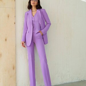 May include: A three-piece lavender suit, consisting of a blazer, vest, and trousers. The blazer has a notched lapel and the trousers are straight-legged. The suit is worn with a silver necklace and matching lavender shoes. The background is a textured white wall.