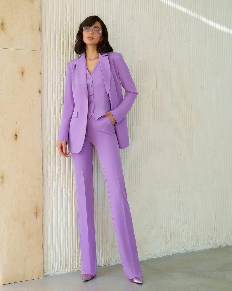 May include: A three-piece lavender suit, consisting of a blazer, vest, and trousers. The blazer has a notched lapel and the trousers are straight-legged. The suit is worn with a silver necklace and matching lavender shoes. The background is a textured white wall.