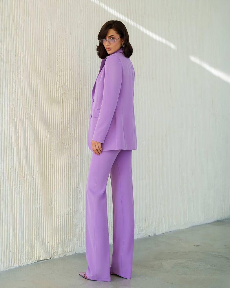 May include: A lavender pantsuit, featuring a double-breasted blazer and wide-leg trousers. The suit is a solid color, with a tailored fit. The person is wearing glasses and heels. The background is a textured white wall.