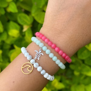 May include: Three bracelets stacked on a wrist. The top bracelet is pink, the middle bracelet is light green, and the bottom bracelet is white with black marble accents. The bracelets have charms, including a silver turtle and a gold wave.