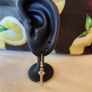 May include: A silver-colored earring shaped like a tower, hanging from a black ear-shaped display stand. The earring has a detailed design, with a hook at the top and a small, star-like base. The background is blurred, with hints of yellow and green.