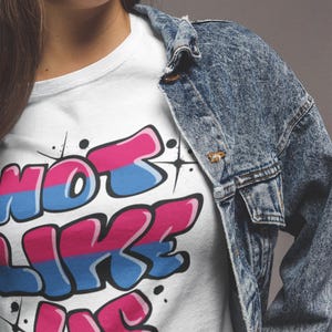 May include: White t-shirt with a graphic print of the words "Not Like Us" in a graffiti style with pink, blue, and white colors.