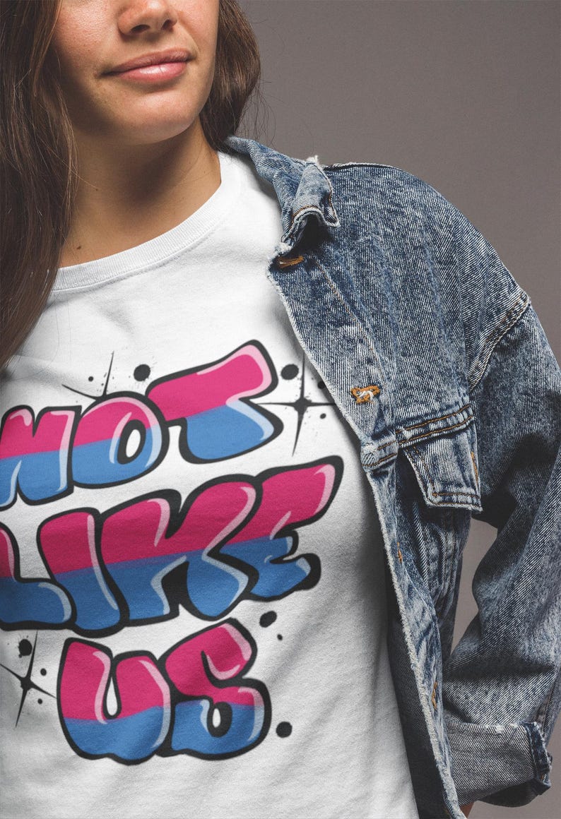 May include: White t-shirt with a graphic print of the words "Not Like Us" in a graffiti style with pink, blue, and white colors.