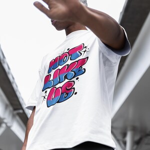 May include: A white t-shirt with a graphic print of the words "NOT LIKE US" in pink, blue, and white. The letters are stylized in a graffiti font.