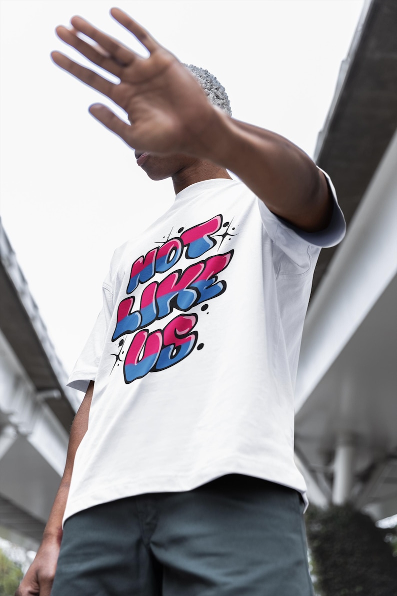May include: A white t-shirt with a graphic print of the words "NOT LIKE US" in pink, blue, and white. The letters are stylized in a graffiti font.