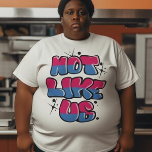 May include: A white t-shirt with a graphic print of the words "NOT LIKE US" in pink, blue, and black bubble letters with white stars.
