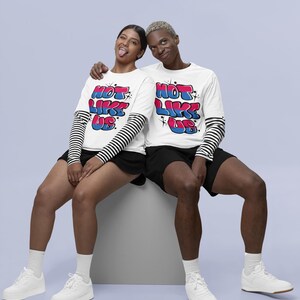 May include: Two people wearing white long-sleeved shirts with the text "Not Like Us" in colorful bubble letters. The shirts have black and white striped sleeves. The people are sitting on a white cube and wearing white sneakers.