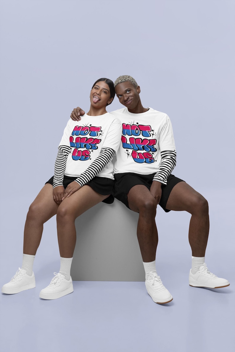May include: Two people wearing white long-sleeved shirts with the text "Not Like Us" in colorful bubble letters. The shirts have black and white striped sleeves. The people are sitting on a white cube and wearing white sneakers.
