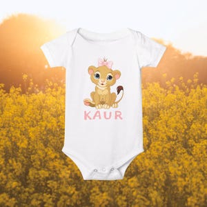 Peut inclure: Combinaison blanche pour bébé avec un lionceau de dessin animé portant un nœud rose. La combinaison a le nom "KAUR" imprimé en lettres roses sous le lion.