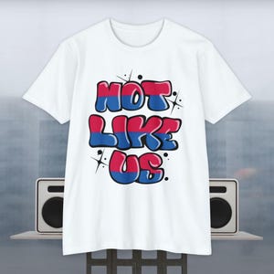 Op de afbeelding: Wit T-shirt met een grafische print van de woorden "Not Like Us" in blauwe en roze bubbelletters met een glittereffect.