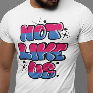 May include: White t-shirt with a graphic print of the words "NOT LIKE US" in pink and blue bubble letters with a white outline. The letters are stylized to look like graffiti.