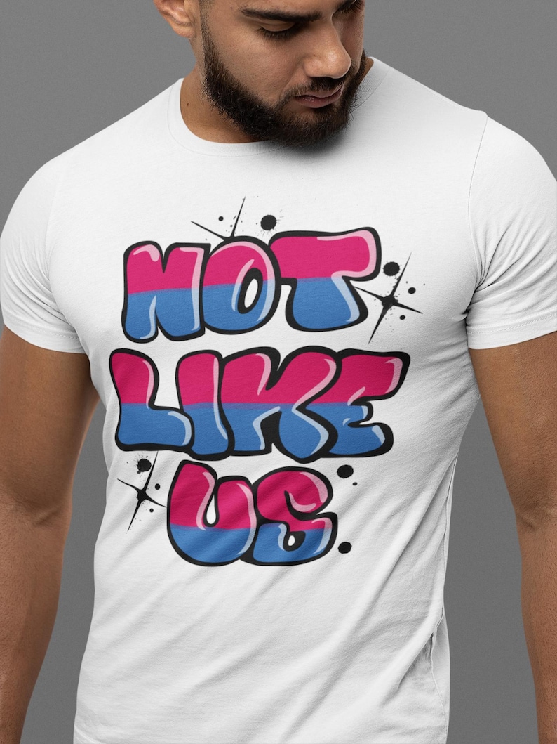 May include: White t-shirt with a graphic print of the words "NOT LIKE US" in pink and blue bubble letters with a white outline. The letters are stylized to look like graffiti.