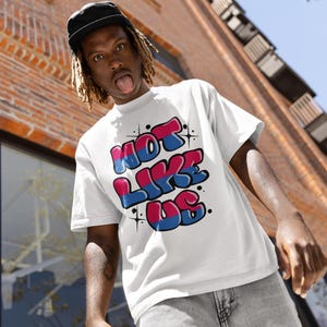 May include: A white t-shirt with a graphic print of the words "NOT LIKE US" in bubble letters with pink and blue accents. The letters are outlined in black.
