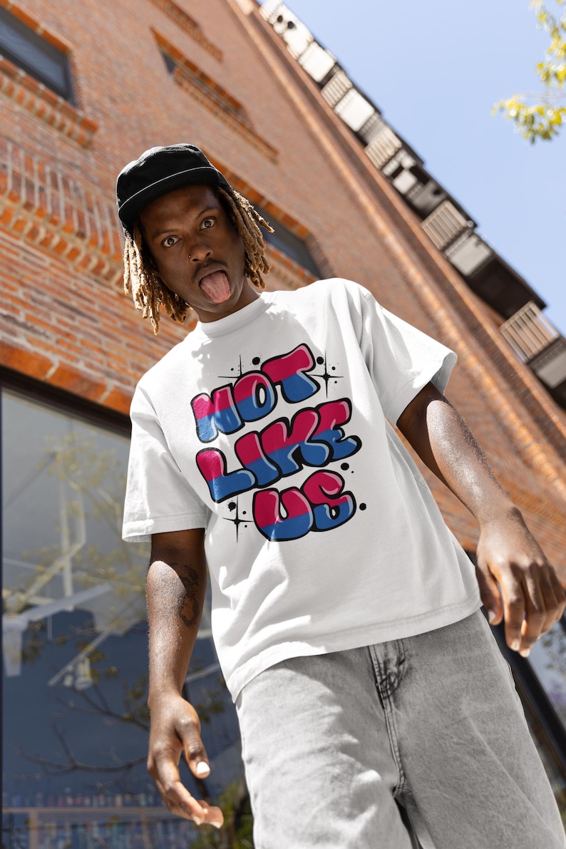 May include: A white t-shirt with a graphic print of the words "NOT LIKE US" in bubble letters with pink and blue accents. The letters are outlined in black.