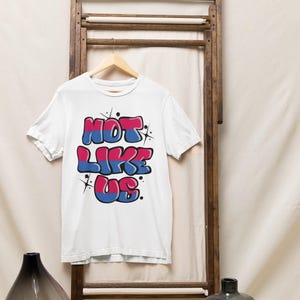 May include: White t-shirt with a graphic print of the text "NOT LIKE US" in pink, blue, and white with a graffiti style. The text is outlined in black and has a glittery effect.