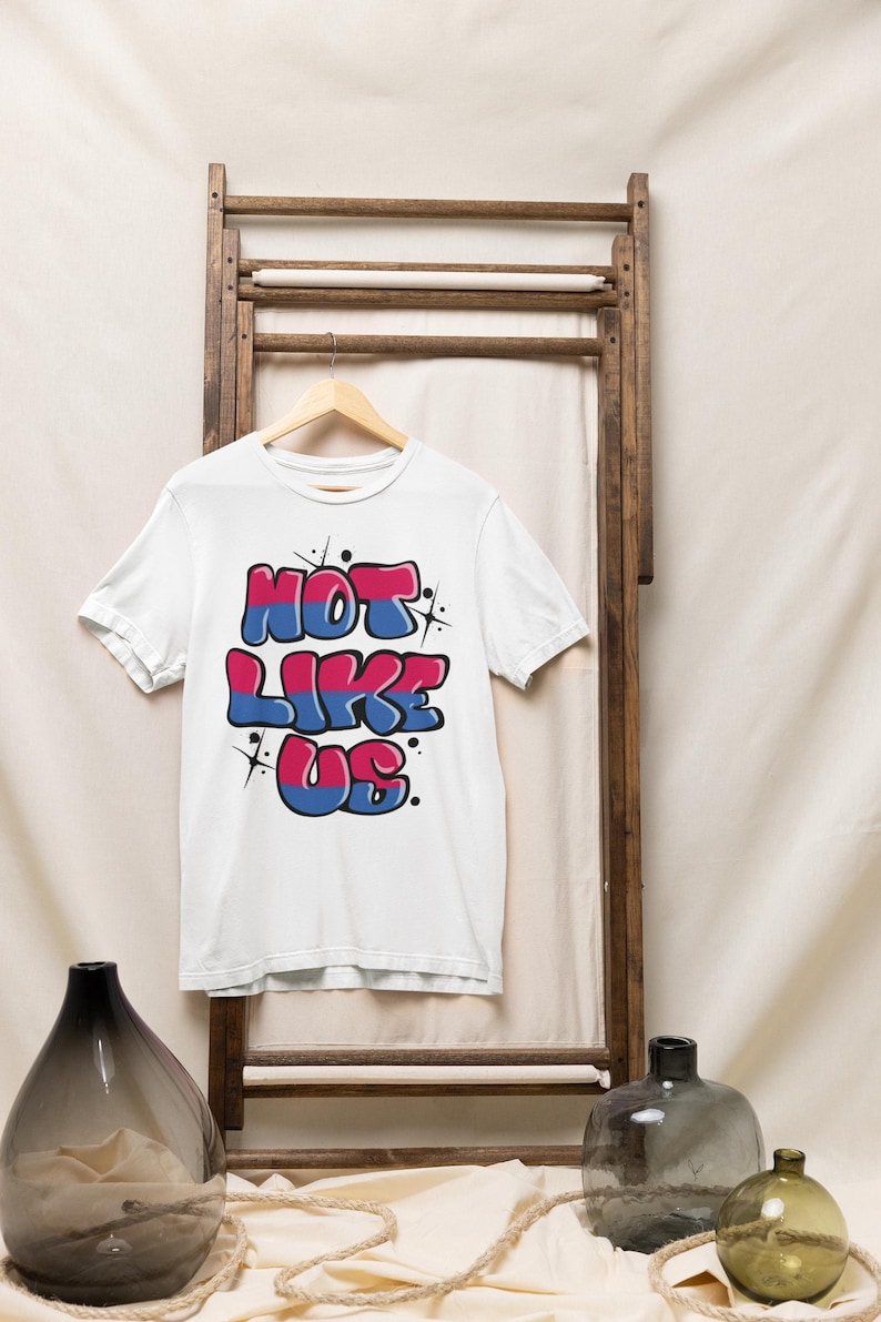 May include: White t-shirt with a graphic print of the text "NOT LIKE US" in pink, blue, and white with a graffiti style. The text is outlined in black and has a glittery effect.
