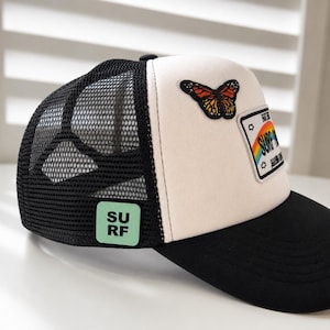 Puede incluir: Gorra de camionero en blanco y negro con una parte trasera de malla y una visera negra. El panel frontal es blanco con un parche de mariposa y un parche que dice "SURF" con un diseño de arcoíris. Un parche cuadrado verde con las letras "SU RF" está en el lateral.
