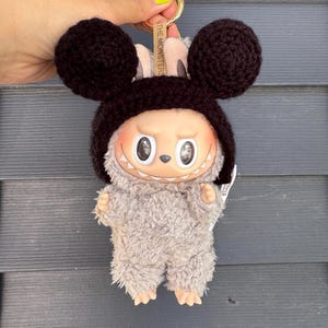 Puede incluir: Llavero de peluche con un personaje con una cara sonriente, ojos grandes y un cuerpo gris difuso. El personaje lleva un gorro de ganchillo marrón oscuro con grandes orejas redondas. El llavero tiene un anillo dorado y una etiqueta que dice "THE MONSTERS".