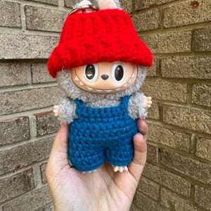 May include: A plush toy with a smiling face, wearing a red crocheted hat and blue overalls. The toy has gray fur and small arms and feet. A gold keychain is attached to the top of the hat.