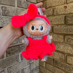 May include: A plush doll with a light brown face, large black eyes, and a wide grin. The doll is wearing a bright red dress and a matching bow. The doll's fur is gray and it has small arms and feet.