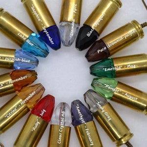 May include: A set of 12 bullet casing earrings with different colored crystal stones. Each earring is labeled with the name of the stone and the month it represents. The stones are: Sapphire, Lake Blue, Periwinkle, Topaz, Citrine, Ruby, Clear Crystal AB, Diamond, Black, Puce, Emerald, and Peridot.