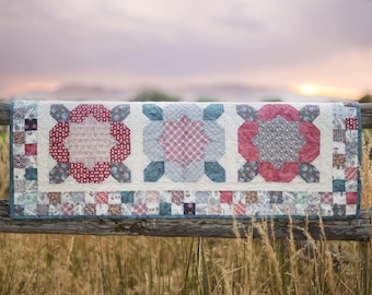 May include: A quilt with a patchwork design featuring three large red, white, and blue star-shaped motifs. The quilt is hanging on a wooden fence against a sunset background.