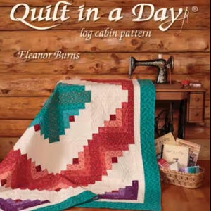 May include: A colorful quilt with a log cabin pattern is draped over a wooden surface. The quilt features red, white, teal, and purple blocks. The book "make a Quilt in a Day" is visible, along with a sewing machine and a basket of sewing supplies.
