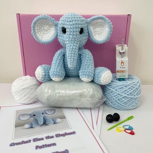 Może przedstawiać: Zestaw do szydełkowania niebieskiego słonia. Zestaw zawiera włóczkę, wypełnienie, szydełko i instrukcje. Zestaw jest zapakowany w różowe pudełko z nadrukiem "Crochet Elsa the Elephant Pattern" na przodzie.