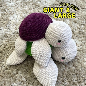 Puede incluir: Un juguete de tortuga de crochet con una concha morada y un cuerpo blanco. La tortuga tiene una parte inferior verde y está sentada en una alfombra marrón. El texto "LOW SS W GIANT & LARGE" es visible en la imagen.